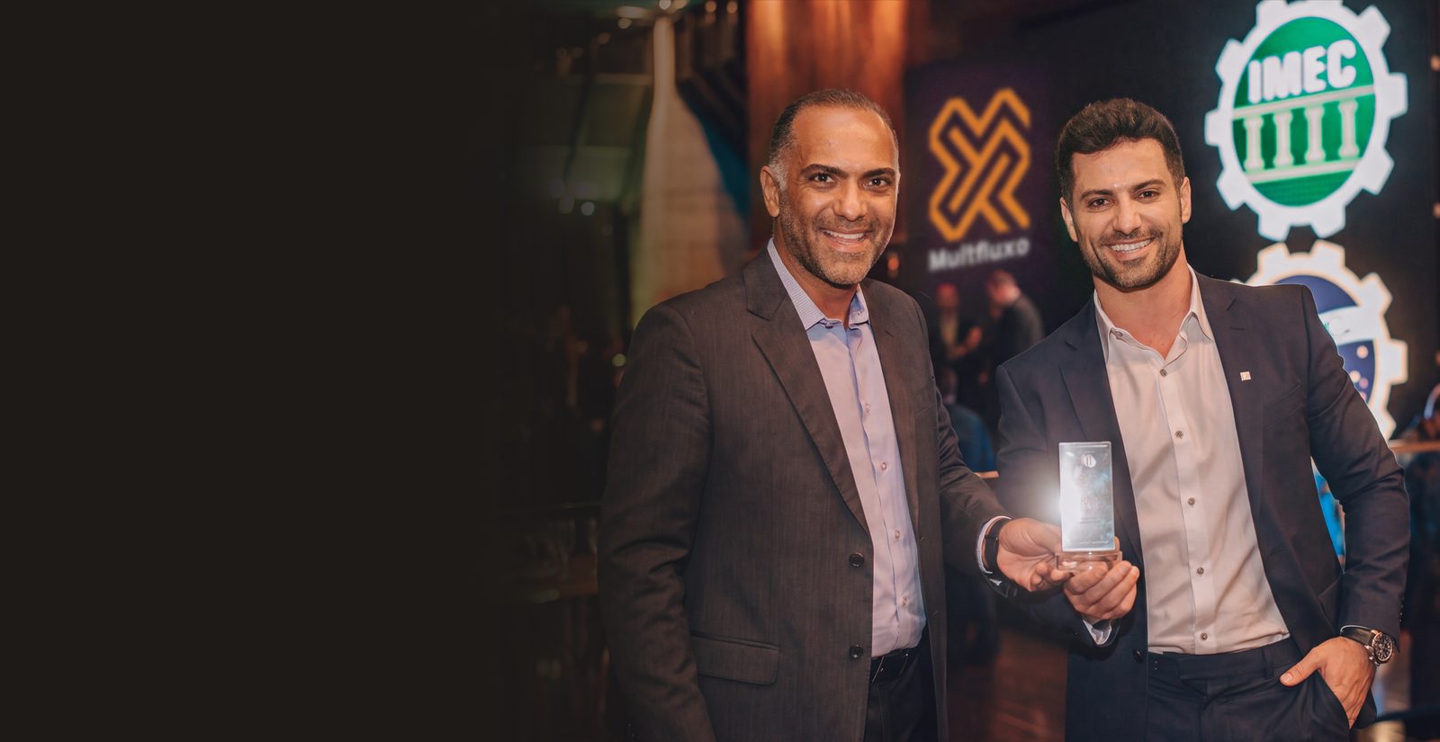 Dois sócios da Multfluxo com o troféu do 18º Prêmio Fornecedores Destaque da Construção Civil MG 2022, diante do painel com as logos da Multfluxo e do IMEC